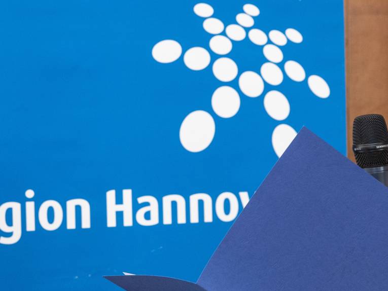Jemand liest aus einer aufgeschlagenen Mappe und spricht in ein Mikrofon. Im Hintergrund ist das Logo der Region Hannover.