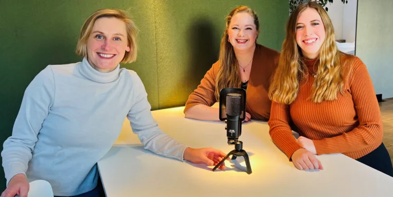 Drei Frauen sitzen an einem Tisch und schauen in die Kamera, auf dem Tisch steht ein Mikrofon.