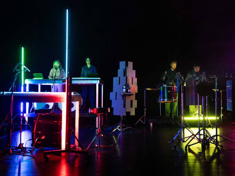 Vier Musiker an Instrumenten auf einer Bühne mit Lichtsäulen