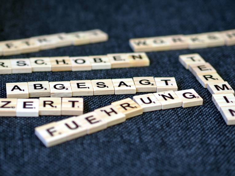 Holzplättchen mit Buchstaben bilden verschiedene Worte. Das Wort "Abgesagt" ist scharf lesbar, während andere Worte unscharf sind.