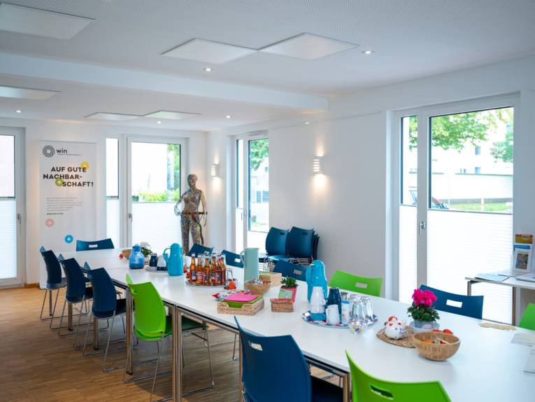 Blick in einen Raum mit einem großen Tisch mit vielen Stühlen. Auf dem Tisch stehen heiße und kalte Getränke, auf einem Rollup-Display steht der Text "AUF GUTE NACHBARSCHAFT".