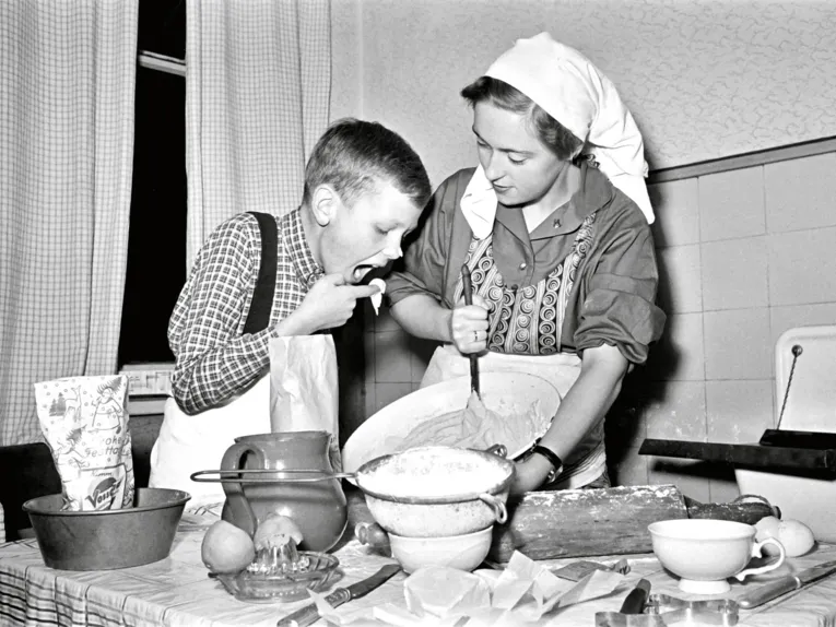 Schwarz-Wei&szlig;-Fotografie: Eine Frau tr&auml;gt Kopftuch und Sch&uuml;rze und r&uuml;hrt mit der Hand einen Kuchenteig. Neben ihr ist ein Junge, der mit dem Finger Teig probiert, der Junge hat auch Kuchenteig an der Nasenspitze.