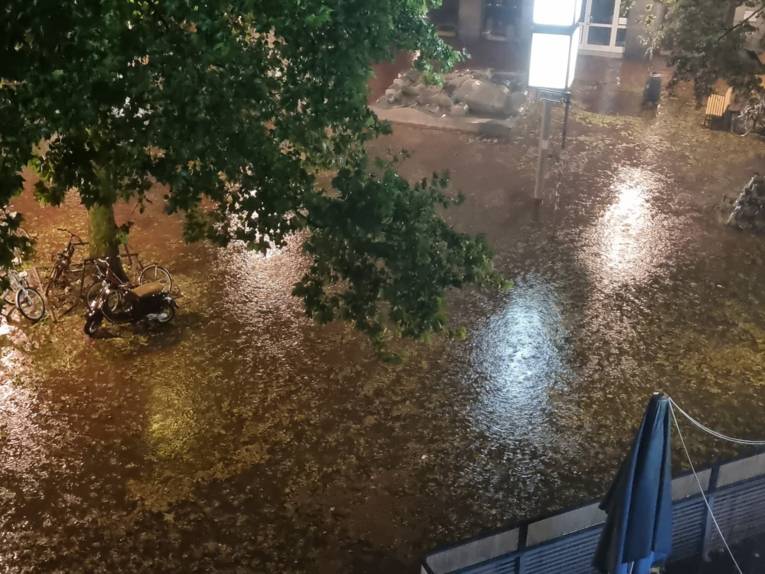 Blick von oben aus einem Gebäude auf einen braune Wasserfläche. Sie steht auf einem breiten Weg zwischen zwei Gebäudezeilen mit einem Baum.