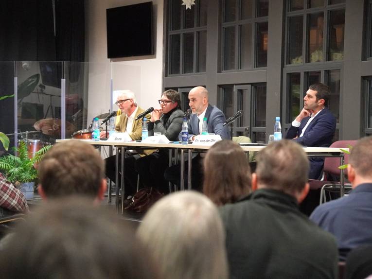 In einem Saal auf einem kleine Pult sitzen eine Frau und drei Männer an Tischen vor Mikrofonen, ein Mann spricht. Vor ihnen sitzen Menschen im Publikum, deren Hinterköpfe zu sehen sind.