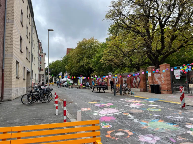 Eine Stra&szlig;e mit Kreidebildern von Blumen, rechts eine lange Girlande aus Luftballons, weiter hinten Tische, B&auml;nke und Zelte.