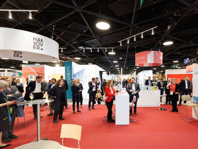 Menschen stehen in einer Halle mit Stehtischen und Ständen.