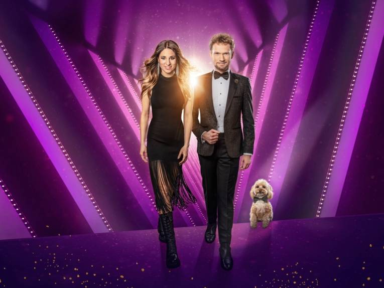 Zu sehen ist das Mentalistenpaar Thommy Ten & Amélie van Tass in formaler Abendbekleidung  vor einem lilafarbenen Showhintergrund.