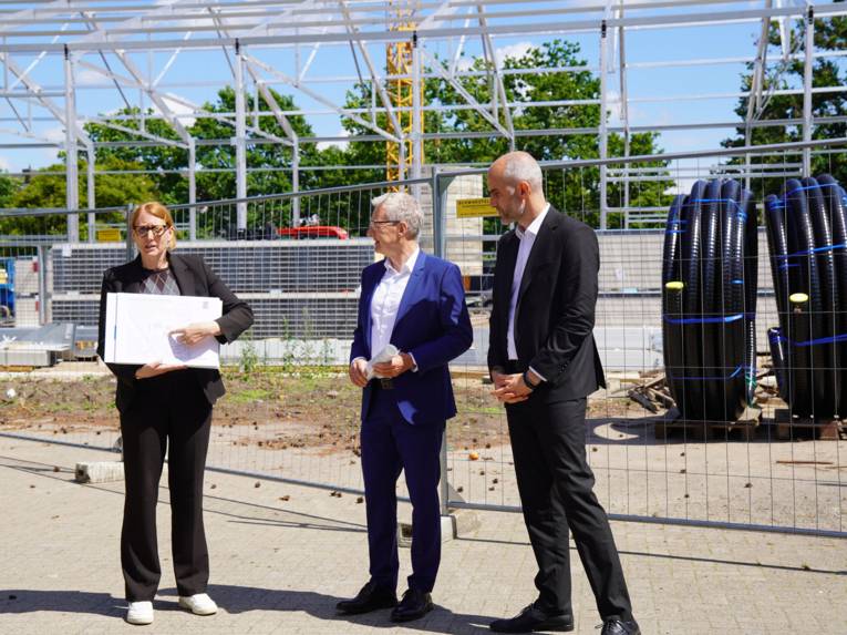Draußen vor einer Baustelle  mit einem großen Metalgerüst vor blauem Himmel stehen eine Frau und zwei Männer. Die Frau hält ein großes Papier und zeigt darauf.