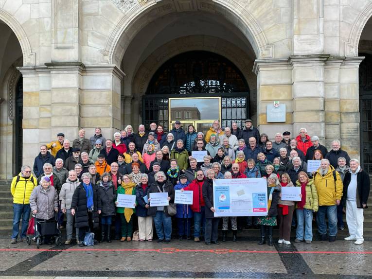 Eine große Gruppe älterer Menschen steht vor und auf einer breiten Treppe vor einem Gebäude mit Bögen über dem Portal, einzele halten Schilder hoch, weitere ein Plakat.
