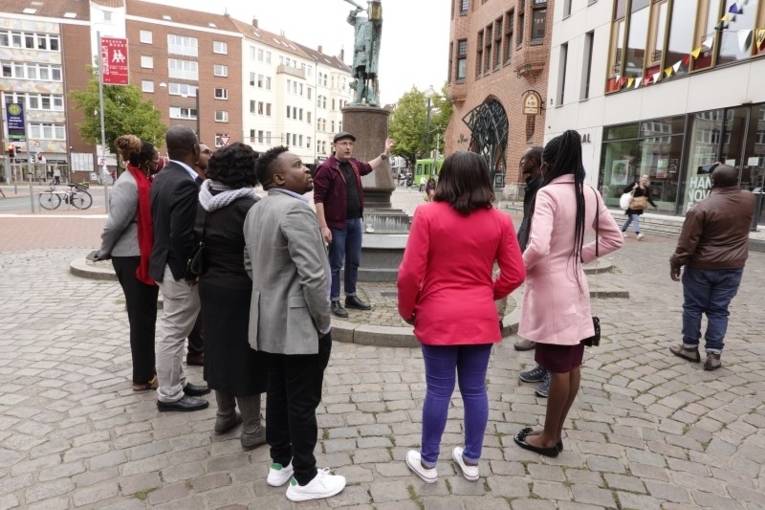 Eine Gruppe Personen auf einem  Platz mit einem Denkmal. Eine weitere Person erklärt etwas.