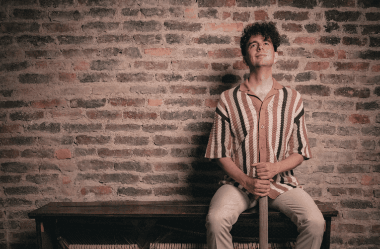 Mann mit kurzen Locken sitzt auf einer Bank vor einer alten Backsteinmauer.