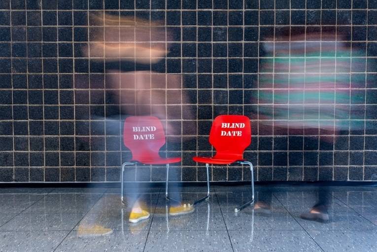 Zwei rote Stühle mit der Aufschrift "Blind Date" vor schwarzem Hintergrund. Davor zwei verschwommene Personen