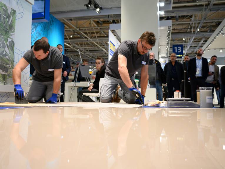 Handwerker verlegen einen Fußboden vor Publikum in einer Messehalle.