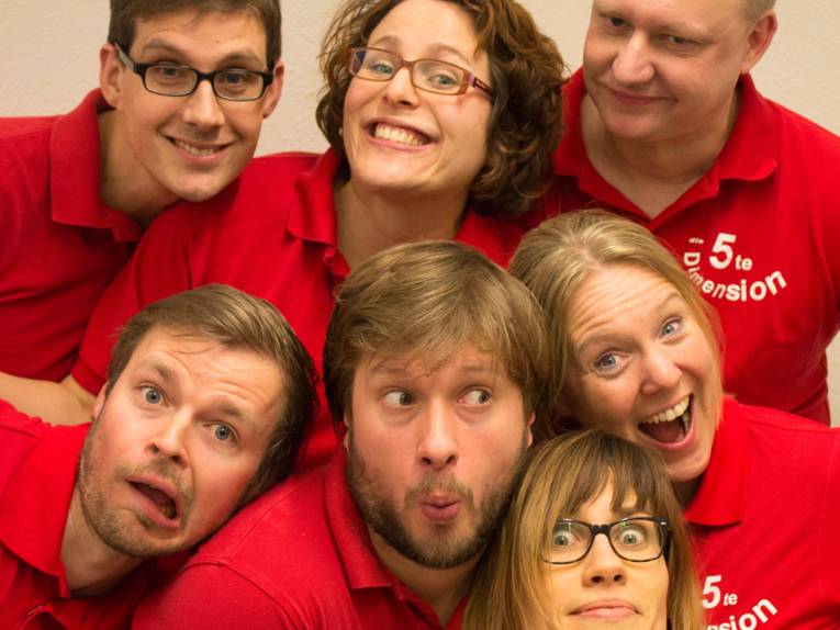 Sieben Menschen, drei Frauen und vier Männer, in roten Polohemden schauen in die Kamera und ziehen dabei Grimassen. Auf einigen der Polohemden ist der Aufdruck „Die 5te Dimension“ zu erkennen.