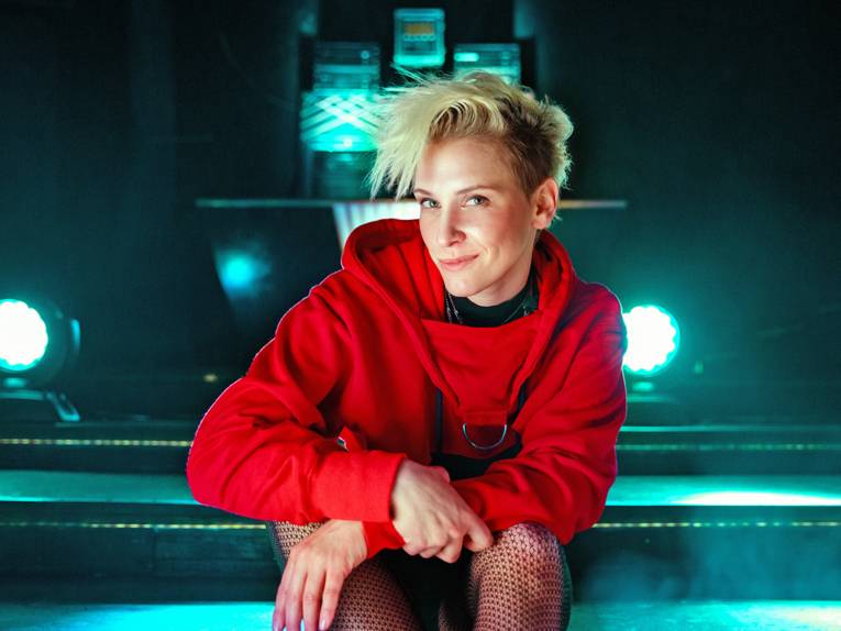 Frau mit kurzen blonden Haaren, trägt ein rotes Oberteil sowie eine Netzstrumpfhose und sitzt auf einer Bühne.