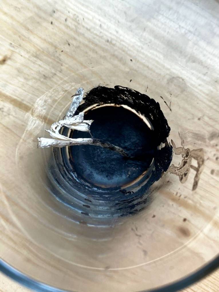 Die Reste eines Pilz sind in einem Glas getrocknet, eine schwarze Substanz hat sich auf dem Glasboden gesammelt.