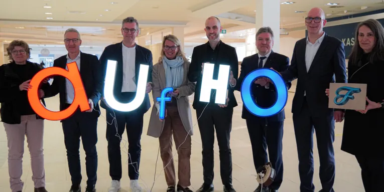 Acht Personen, die leuchtende Buchstaben halten. Diese bilden das Wort "aufhof".