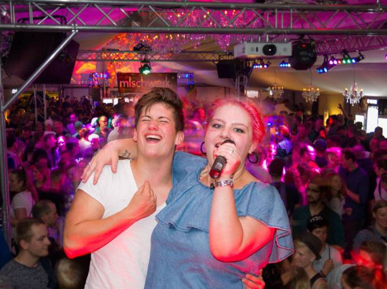 Zwei Personen in einem Zelt, die Karaoke singen. Im Hintergrund unz&auml;hlige Menschen.