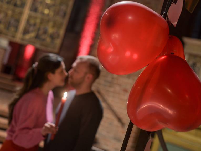 Eine Frau und ein Mann halten in einer Kirche eine Kerze und küssen sich, im Vordergrund hängen Herz-Luftballons.