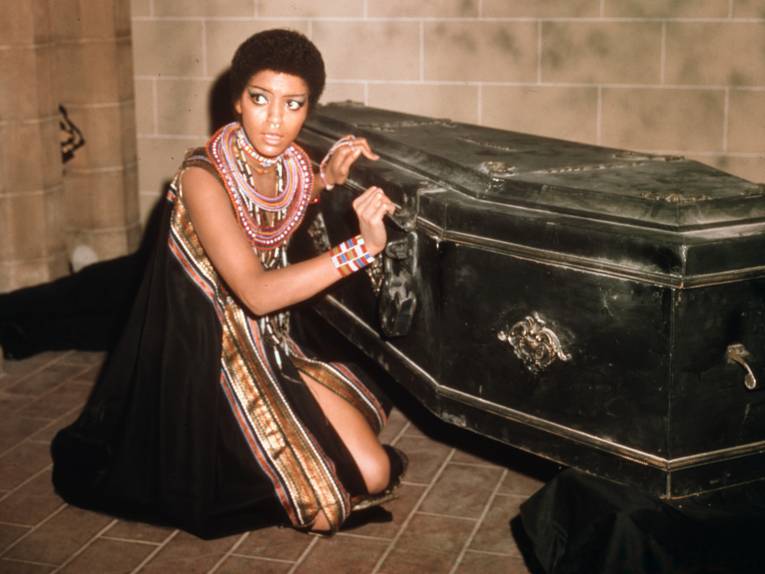 Eine Frau kniet vor einem schwarzen Sarg. Es sieht aus, als wolle sie diesen öffnen.