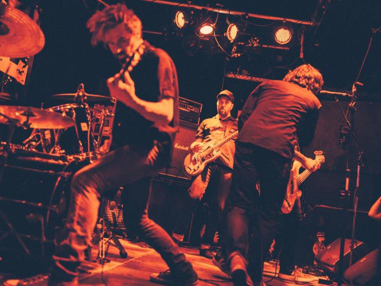 Drei Männer mit E-Gitarren und ein Mann mit Mikrofon auf einer Bühne.