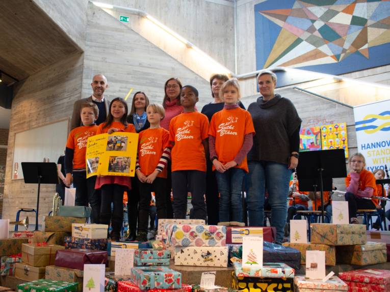 Fünf Kinder mit orange-farbenen T-Shirts und fünf Erwachsene stehen, von Geschenken umgeben, in einer Schulhalle.