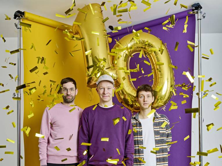 Drei Männer stehen vor einer lila-gelben Wand im Konfetti-Regen, zudem hängt eine goldene Luftballon-Zehn über ihnen.