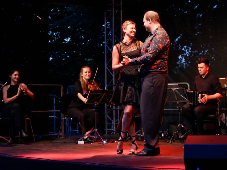 Ein Tänzer und eine Tänzerin tanzen Tango auf einer Bühne, im Hintergrund spielen Musikerinnen und Musiker.