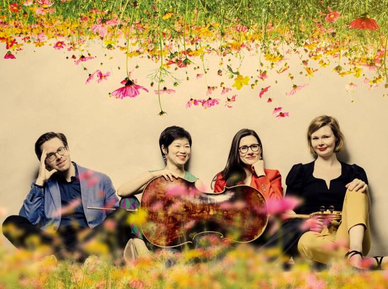 Drei Frauen und ein Mann sitzen hinter einem querliegenden Cello auf einer Blumenwiese, von oben wachsen weitere Blumen ins Bild hinein.