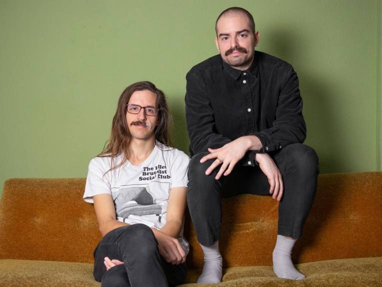 Ein Mann mit langem Haar und Brille sitzt auf einem Sofa, auf der Lehne ein zweiter mit sehr kurzem Resthaar und Schnauzbart.