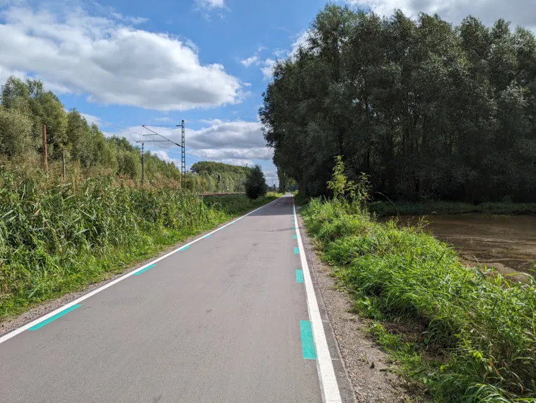 Ein asphaltierter Weg, links gr&uuml;ner Bewuchs, daneben Bahnschienen und Oberleitungen, rechts vom Weg gr&uuml;ner Bewuchs und B&auml;ume.