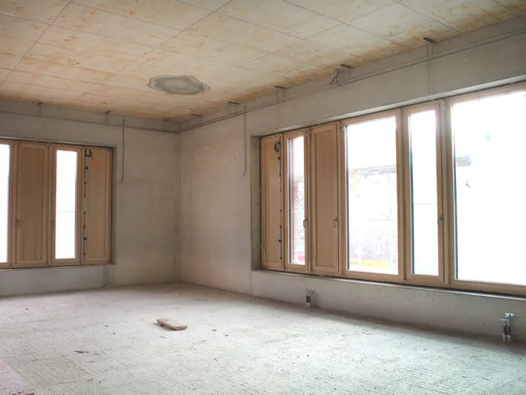 Die Ecke eines Raums in Beton und Estrich, links hinten und rechts hohe Fensterfronten.