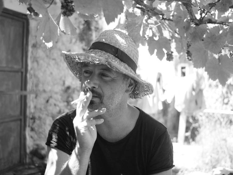 Zu sehen ist ein schwarzweißes Foto eines mittelalten Mannes, der in einem sonnigen Innenhof sitzt und eine Zigarette raucht.