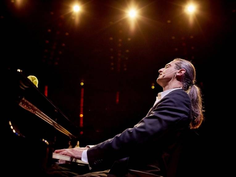 Zu sehen ist ein Mann mit Pferdeschwanz, der am Klavier sitzt.