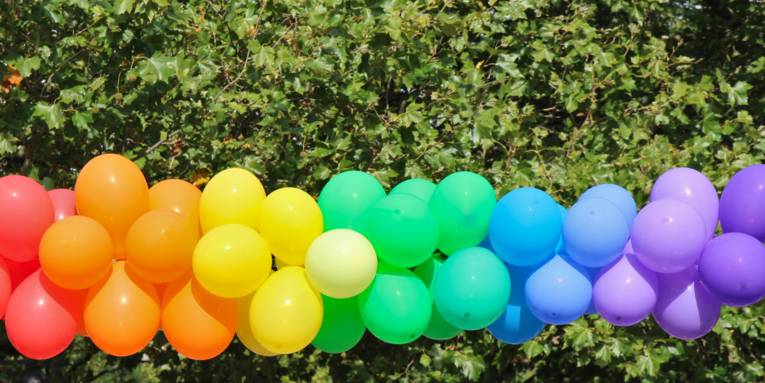 Luftballons sind draußen bei einer Veranstaltung so aufgehängt, dass sie einen Farbverlauf wie bei einem Regenbogen bilden.