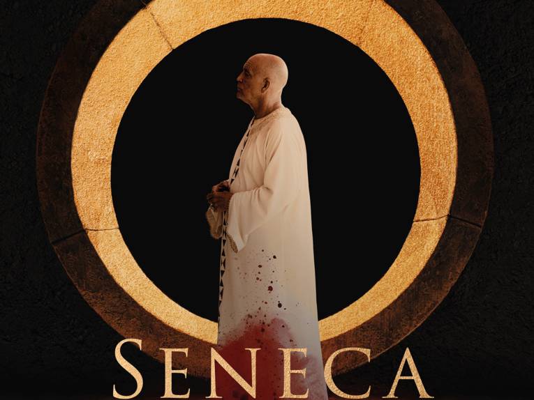 Zu sehen ist eine ältere Person in einem weißen Gewand, die in einem goldenen Kreis steht. Der Schriftzug "Seneca" ist blutbefleckt.