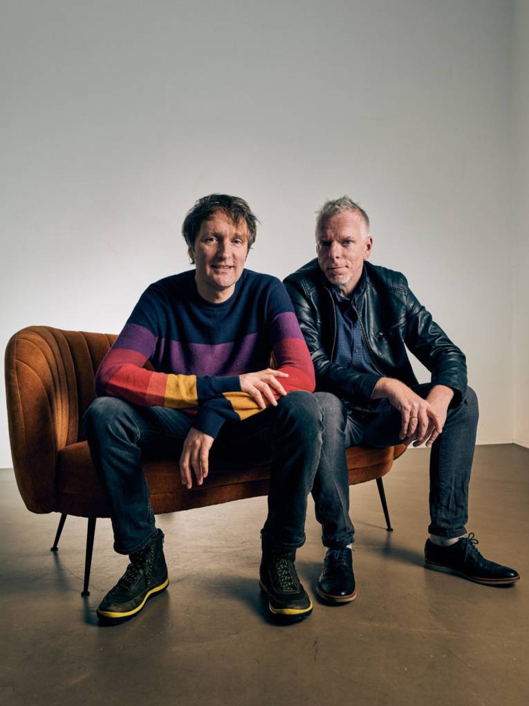 Zwei M&auml;nner sitzen auf einem Polsterm&ouml;belst&uuml;ck und schauen in die Kamera.