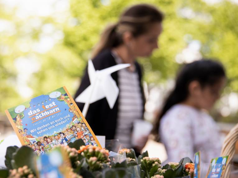 Im Vordergrund ein Flyer "Das Fest der Nachbarn", umgeben von Blumen, im Hintergrund unscharf zwei Personen