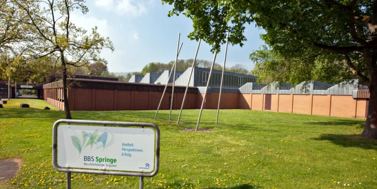Ein Schild steht auf einer Rasenfläche, darauf der Text "BBS Springe. Berufsbildende Schulen. Vielfalt. Perspektiven. Erfolg." Hinter dem Schild ist ein großer Schulgebäudekomplex.