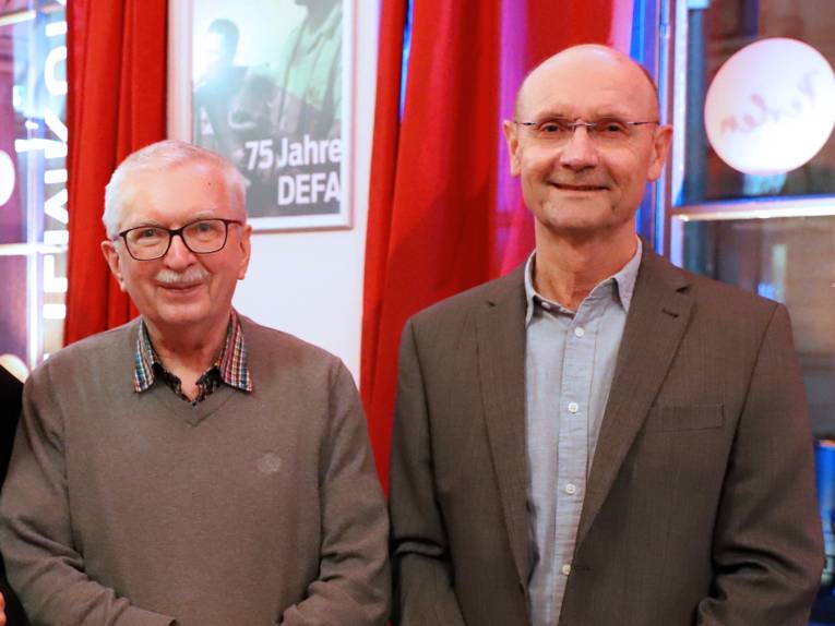 zwei Männer stehen vor einem Plakat, auf dem 75 Jahre DEFA steht