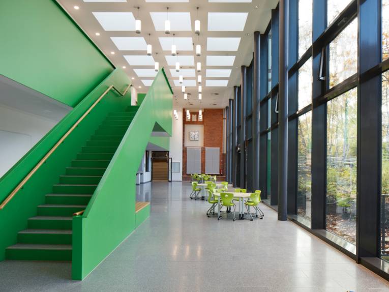 Ein hoher langer Raum, rechts hohe Fenster, links eine Treppe mit gr&uuml;ner Abgrenzung, unten im Raum stehen kleine Tische mit gr&uuml;nen St&uuml;hlen.