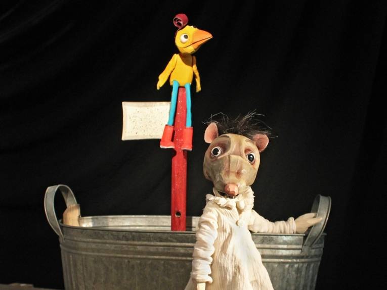 Zu sehen ist eine Theaterpuppe: Eine Ratte steht vor einem Bottich, das durch aufgrund eines roten Mastes an ein Boot erinnert. Oben auf dem Mast sitzt eine gelbe Vogelfigur.