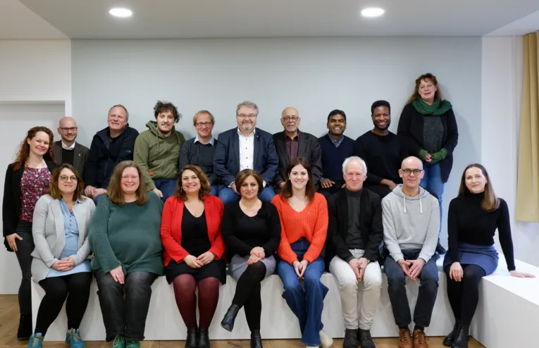 18 Personen sitzen in zwei Reihen hintereinander auf Podesten. Sie sind zu einem Gruppenbild positioniert und lächeln in die Kamera.