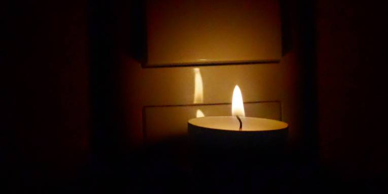 Ein Teelicht steht auf dem massiven Stecker einer LED-Lampe vor dem Lichtschalter einer Deckenbeleuchtung. Nur die Flamme bringt etwas Licht, drumherum ist es dunkel.