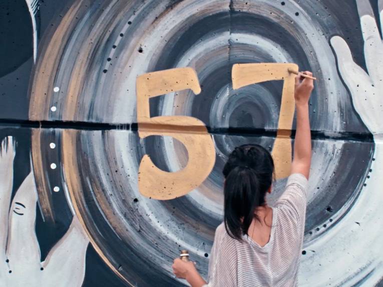 Eine Person mit Pferdeschwanz malt an eine Wand die Zahl 57