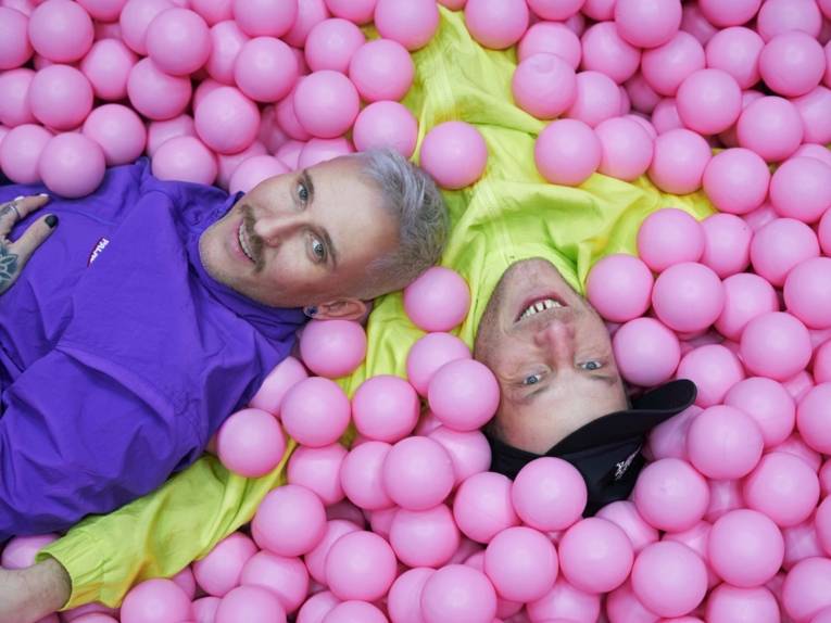 Zwei Männer liegen in einem rosafarbenen Bällebad.