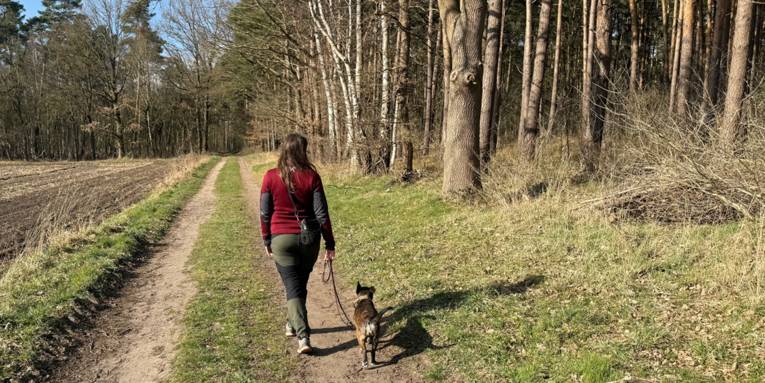 Eine Frau f&uuml;hrt einen Hund an der Leine auf einem Weg zwischen Feld und Wald.
