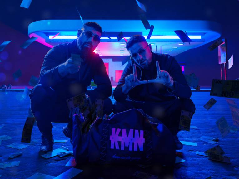 Zwei Männer hocken auf dem Boden, hinter ihnen blaues Neonlicht.