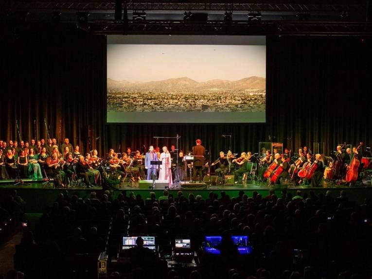 Zu sehen ist ein Orchester vor einer großen und hoch hängenden Leinwand auf einer Bühne.