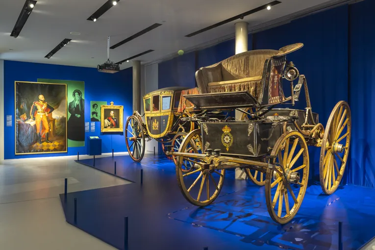 Blick in die Ausstellung "Vier Kutschen, ein Königreich. Hannover 1814 - 1866" , im Vordergrund ein Phaeton, dahinter ein Reisewagen, an der Wand Gemälde von Herrscher und ihren Ehefrauen.